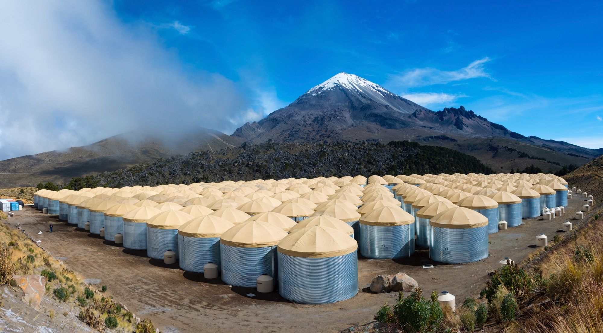 Los tanques de agua del observatorio HAWC, cilindros de acero con techo de lona dispuestos en una parrilla compacta en la ladera del volcán, con montañas al fondo.